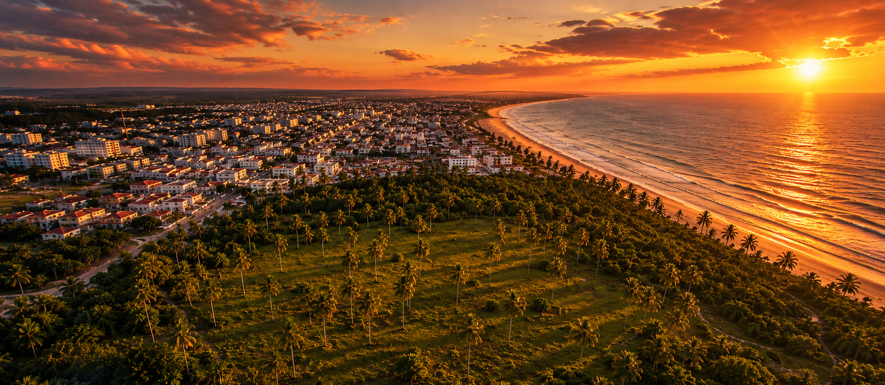 Luftaufnahme eines Küstenortes mit Strand, Palmen und angrenzendem Entwicklungsareal bei Sonnenuntergang